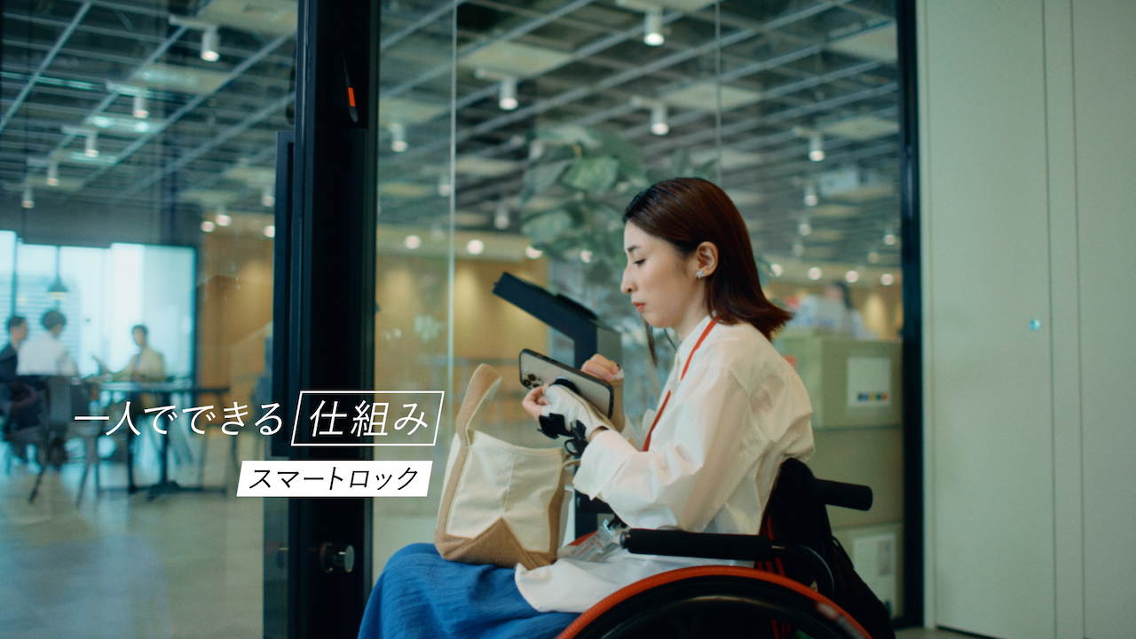 ドアの前でスマートフォンを操作する車椅子の女性と「一人でできる仕組み スマートロック」のテロップ
