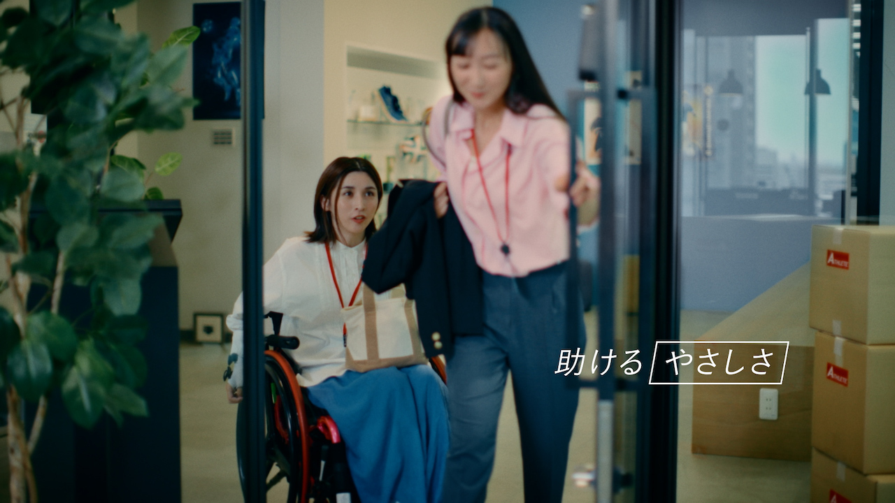 車椅子の女性に代わって、ドアを開ける同僚の女性と「助けるやさしさ」のテロップ